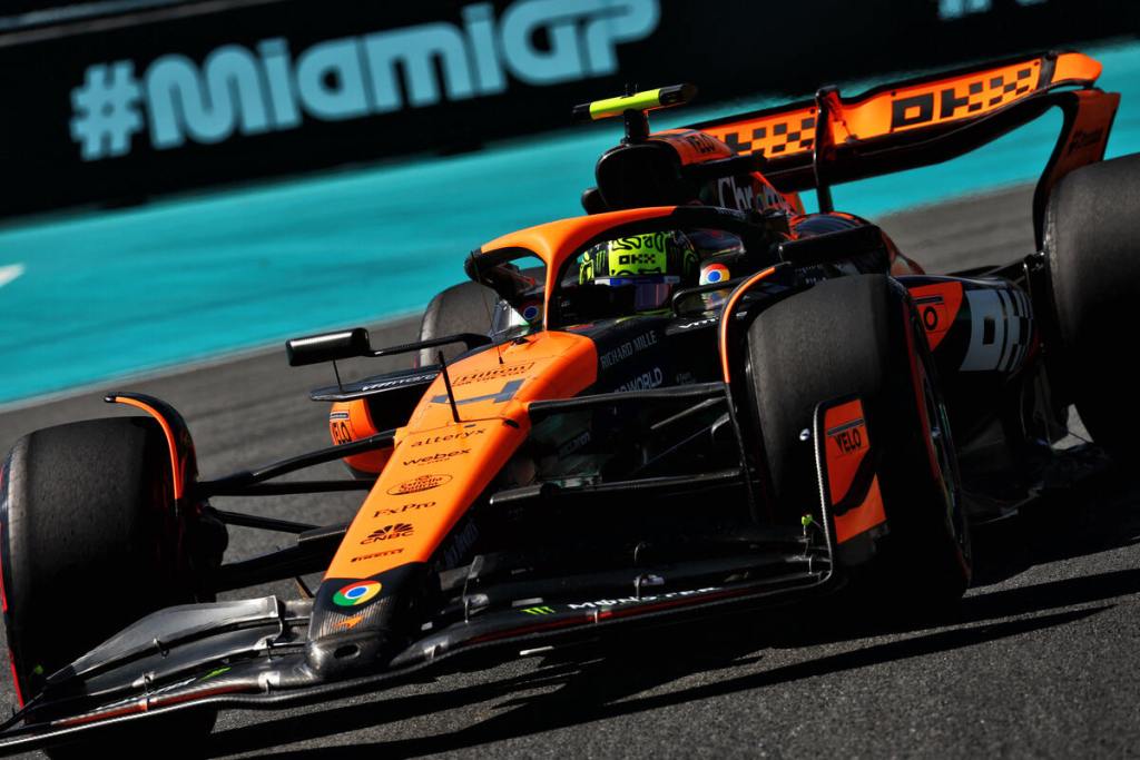Una McLaren Formula 1 di colore arancione in movimento sul circuito di Miami, con il logo del Gran Premio di Miami sullo sfondo.