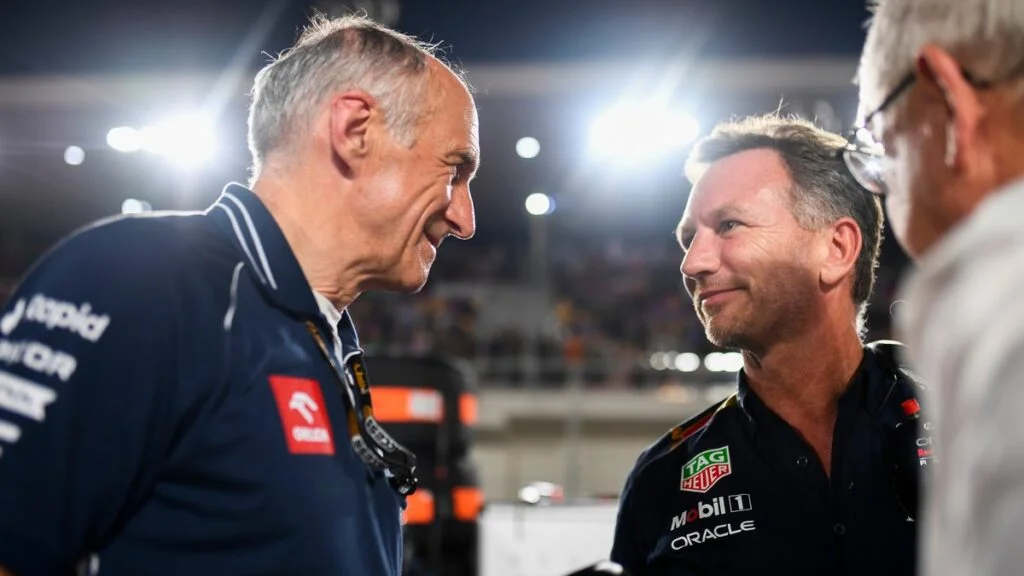 Toast e Horner discutono a un evento di Formula 1, con sorrisi e interazioni amichevoli, sullo sfondo di un ambiente illuminato e affollato.